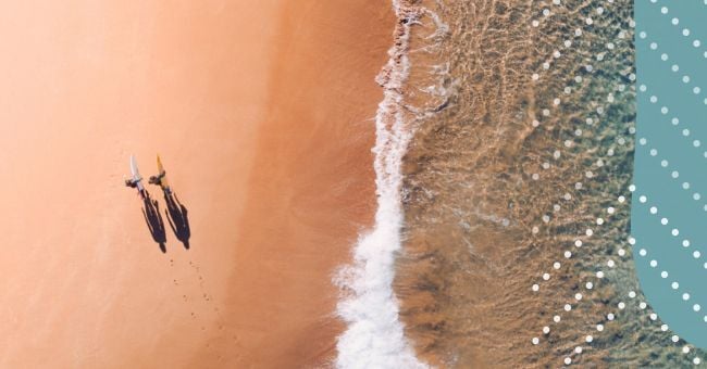 Two people walk on a beach next to waves seen from above