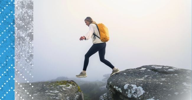 A hiker dressed in black and white steps from one rock to another high up on a montainside in the fog.