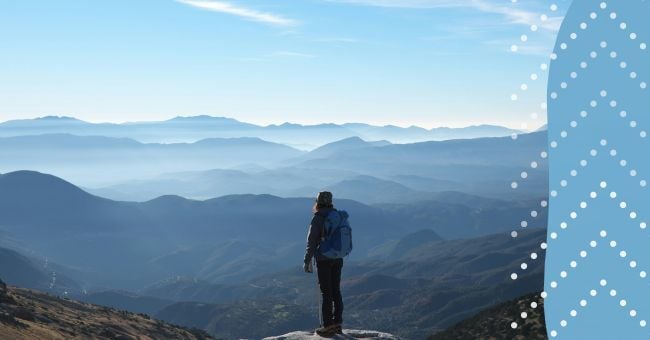 A person stands on a mountain looking at valleys and hills in the distance