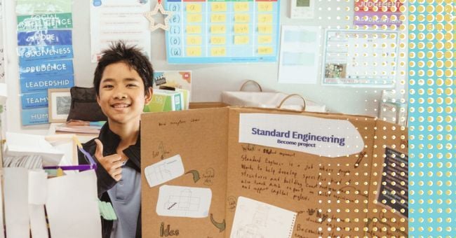 A year 7 student smiles proudly in front of their project investigating what a Standards Engineer does. They are sitting at a table in a colourful classroom