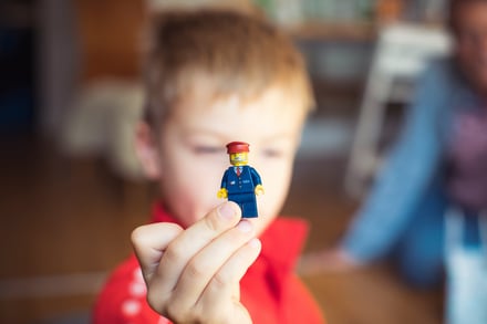 A young child holds a play figure towards the camera. The child is obscured but we see the play figure clearly - it is a person in a hat and uniform such as a train driver or postal worker. 