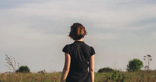 A young woman in a black shirt seen from the back. She is in a grassy field looking at the horizon. There is a thoughtful mood to the photo.