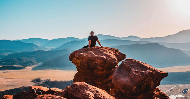 A young person seen from the back sitting on top of a rock looking over a valley at distant mountains