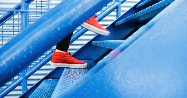 A pair of feet in red shoes climb a blue staircase. 