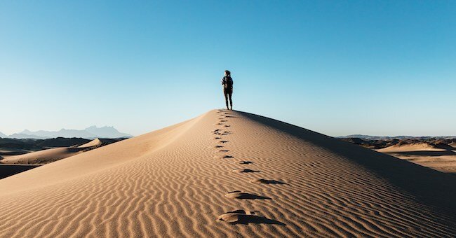 A person exploring - their footsteps leading up a sand dune. Blue sky beyond. 