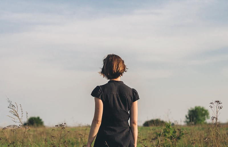 A yougn woman in a black shirt seen from the back. She is in a grassy field looking at the horizon. There is a thoughtful mood to the photo.